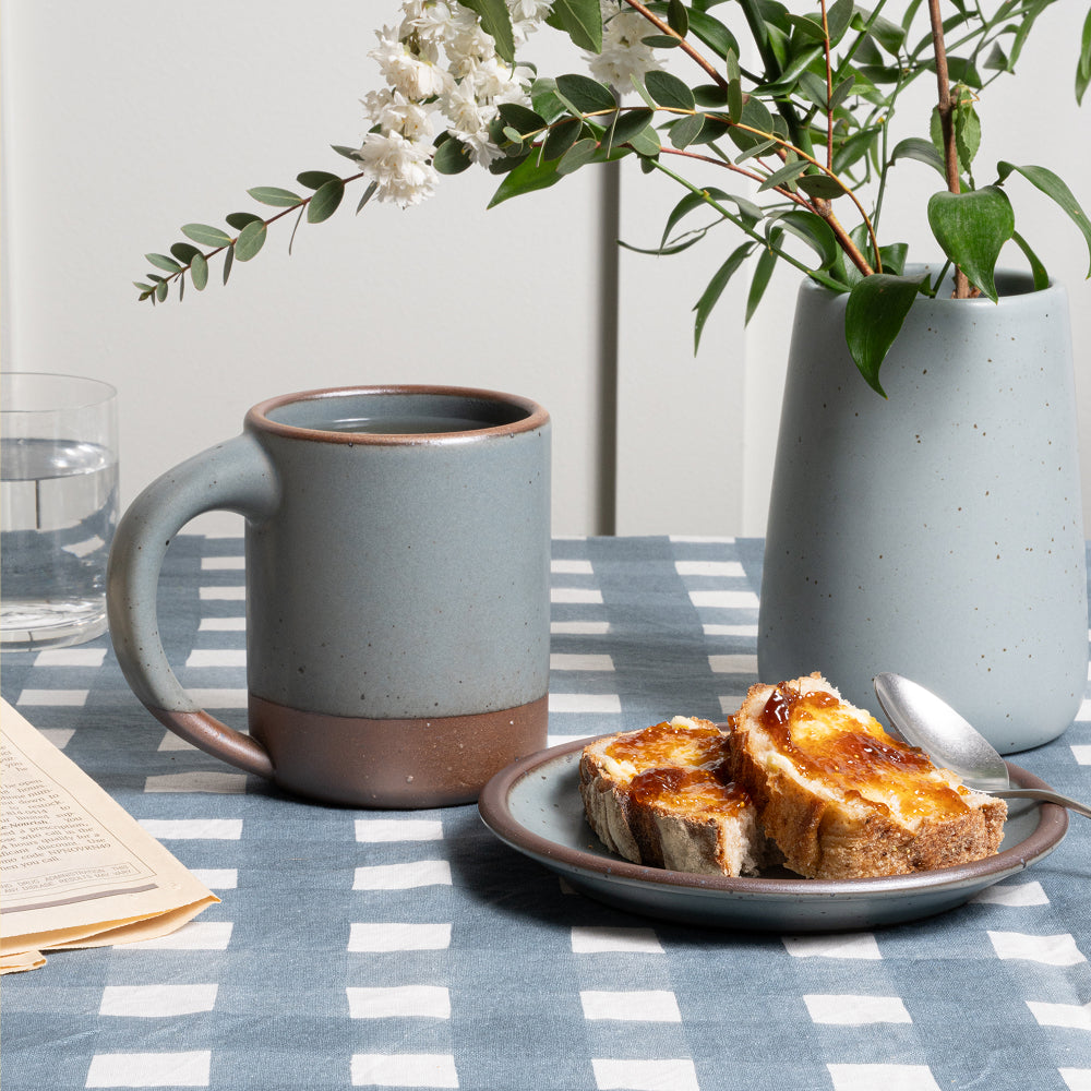 A ceramic mug in a blue-grey color with an unglazed base and rim, along with a matching cake plate and a similar slope vase, all on a blue-grey crosshatch tablecloth.