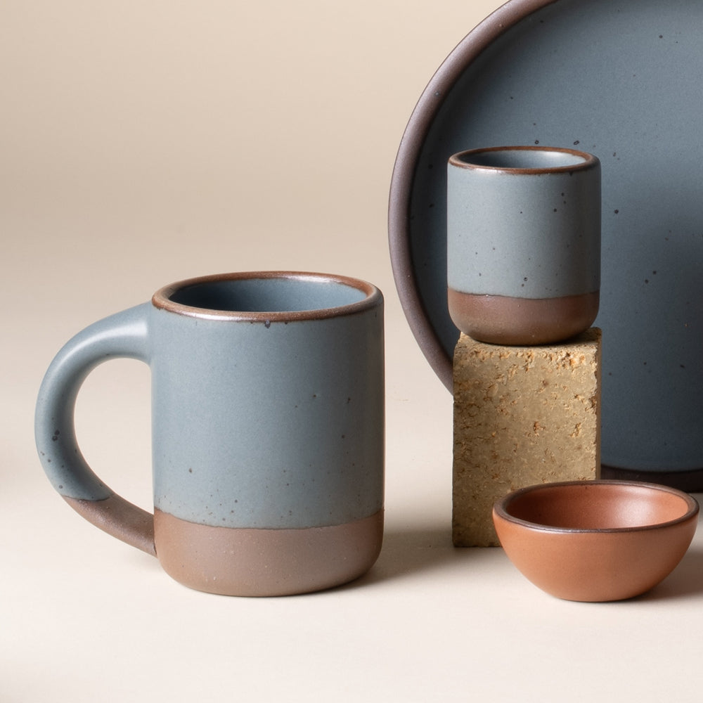 A ceramic mug in a blue-grey color with an unglazed base and rim, next to a tiny cup, along with a small bowl and oval platter.