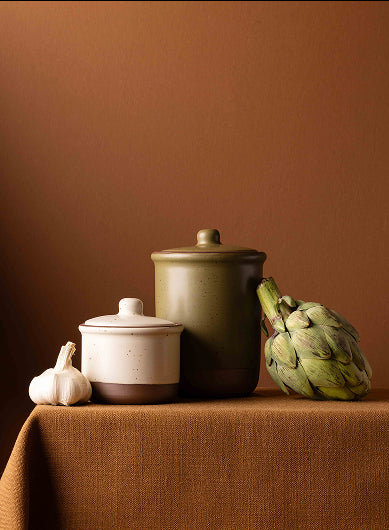 Three ceramic jars with matching lids are arranged on a tablecloth against a warm brown background. The canisters have a two-tone design with a dark brown base and a light beige or cream upper body.