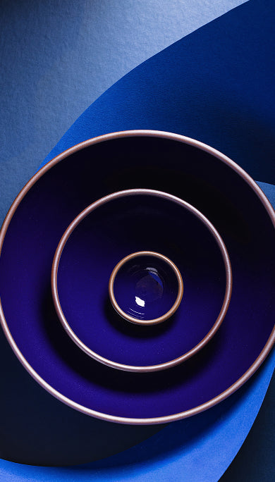 Three nested ceramic bowls in a dark glossy blue with contrasting brown rims, arranged diagonally on a matching blue textured background.