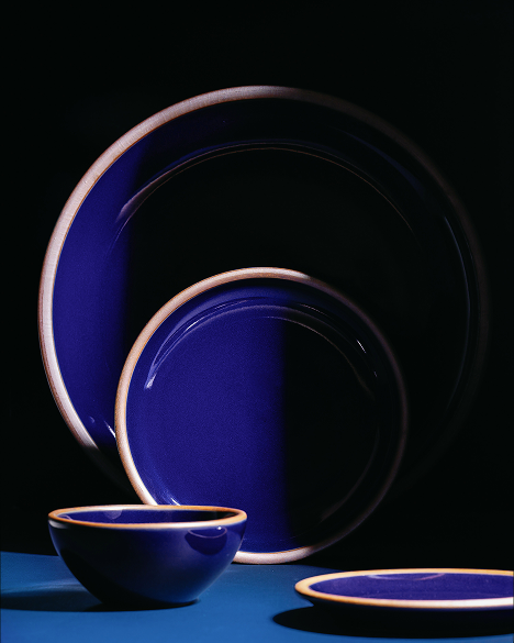 A coordinated set of ceramic plates and a bowl in glossy dark blue with a contrasting light brown rim, stacked in front of a dark background. 