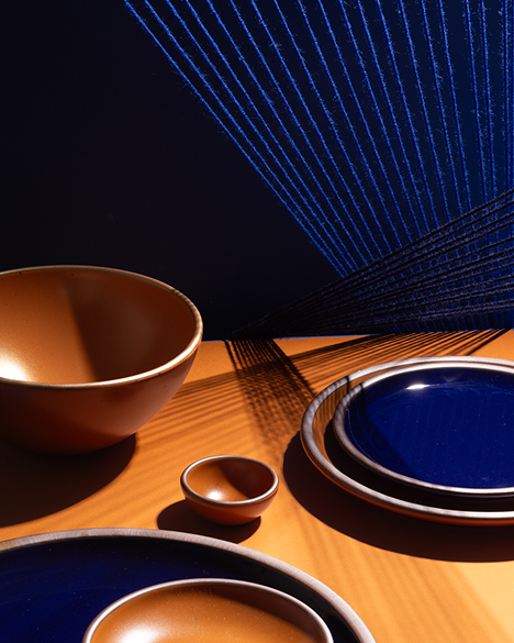 A styled tabletop arrangement of ceramic tableware, including glossy dark blue and earthy orange plates and bowls with light brown rims, set on a warm wood surface under dramatic directional light that casts shadows.