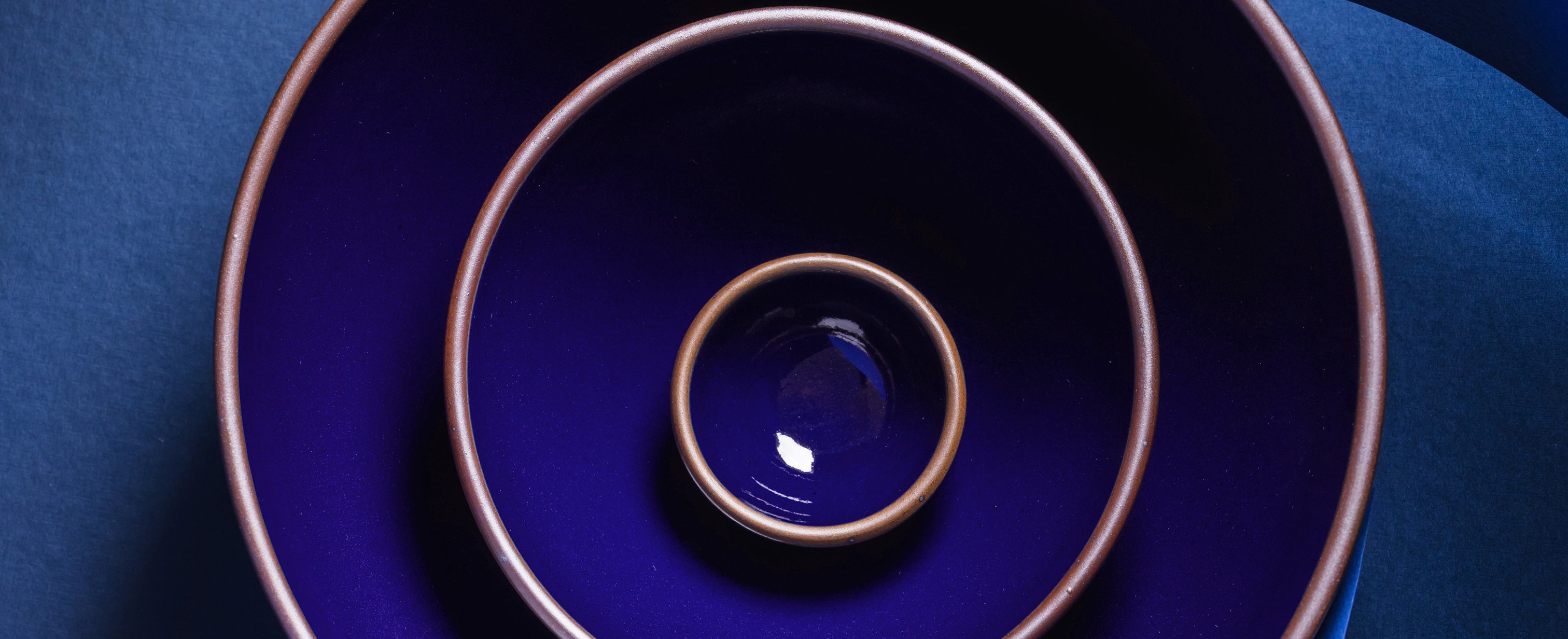Three nested ceramic bowls in a glossy dark blue glaze with a contrasting brown rim, arranged concentrically on a dark blue background.