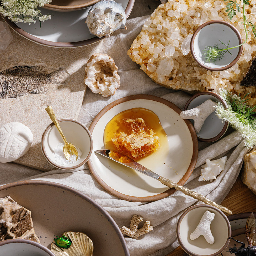 A styled flat-lay of ceramic tableware on a linen cloth, featuring a ceramic dessert plate with honeycomb and honey, surrounded by seashell decor, crystals, and small bowls.