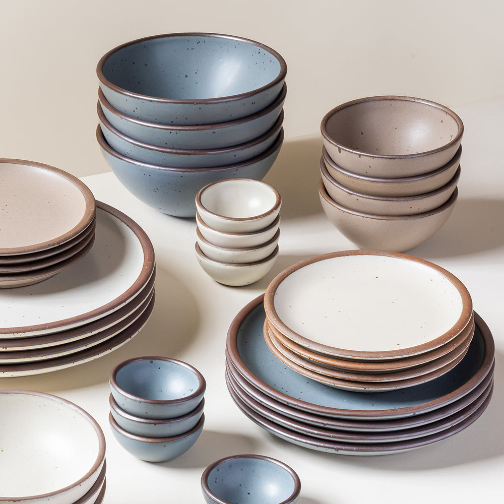 Stacks of ceramic plates and bowls in blue-grey, white, and neutral glazes on a white surface.