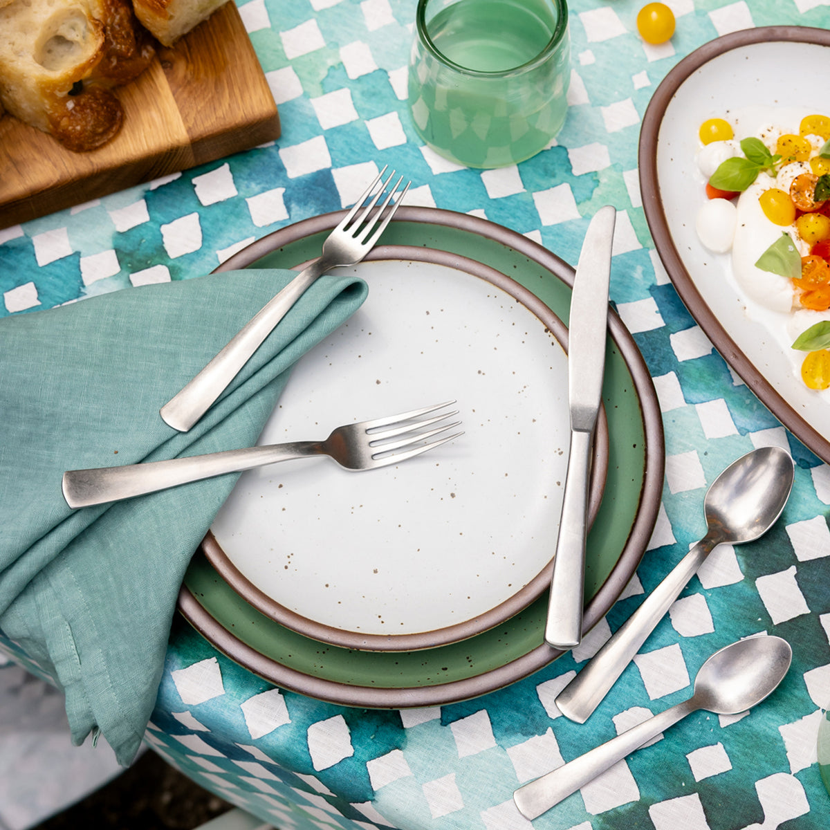 Cool white speckled ceramic plate stacked on a green plate with silver flatware and a teal linen napkin on a blue-and-white patterned tablecloth.