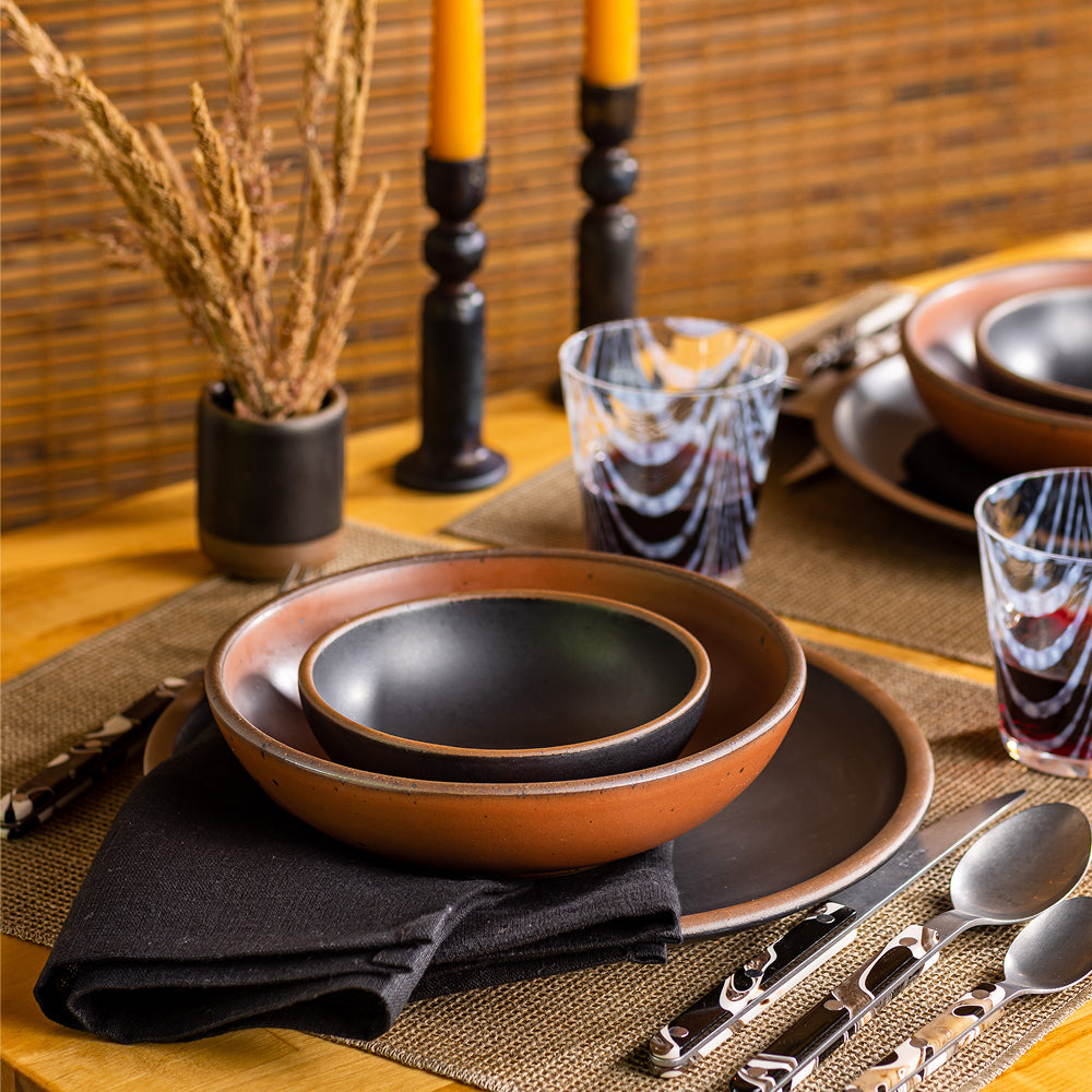 A place setting of ceramic bowls and plate in a black and cool terracotta colors with patterned glassware and marbled flatware.