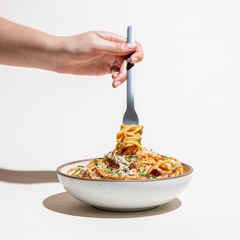 A hand holding a fork above a shallow ceramic bowl of spaghetti with tomato sauce, grated cheese, and green herbs on a clean white background.