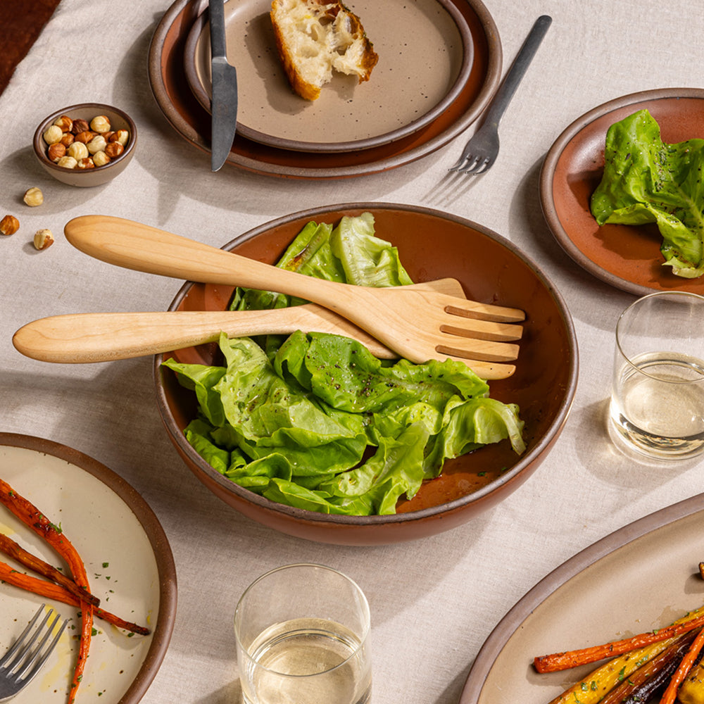 A large shallow ceramic bowl in a cool terracotta color on a table with salad, wood serving set and other plates with food.