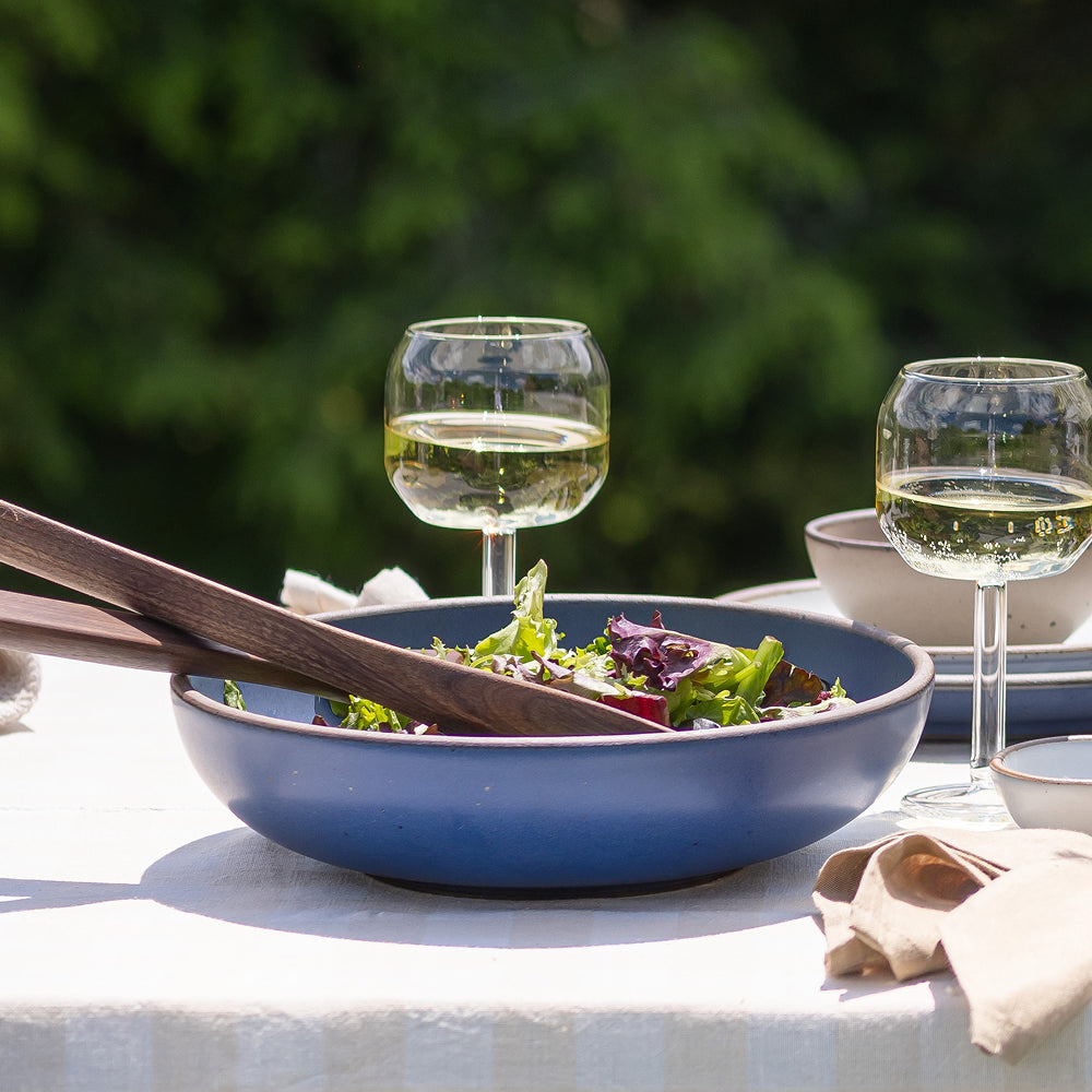 Large shallow blue ceramic serving bowl filled with salad and wooden servers on an outdoor table with two wine glasses.