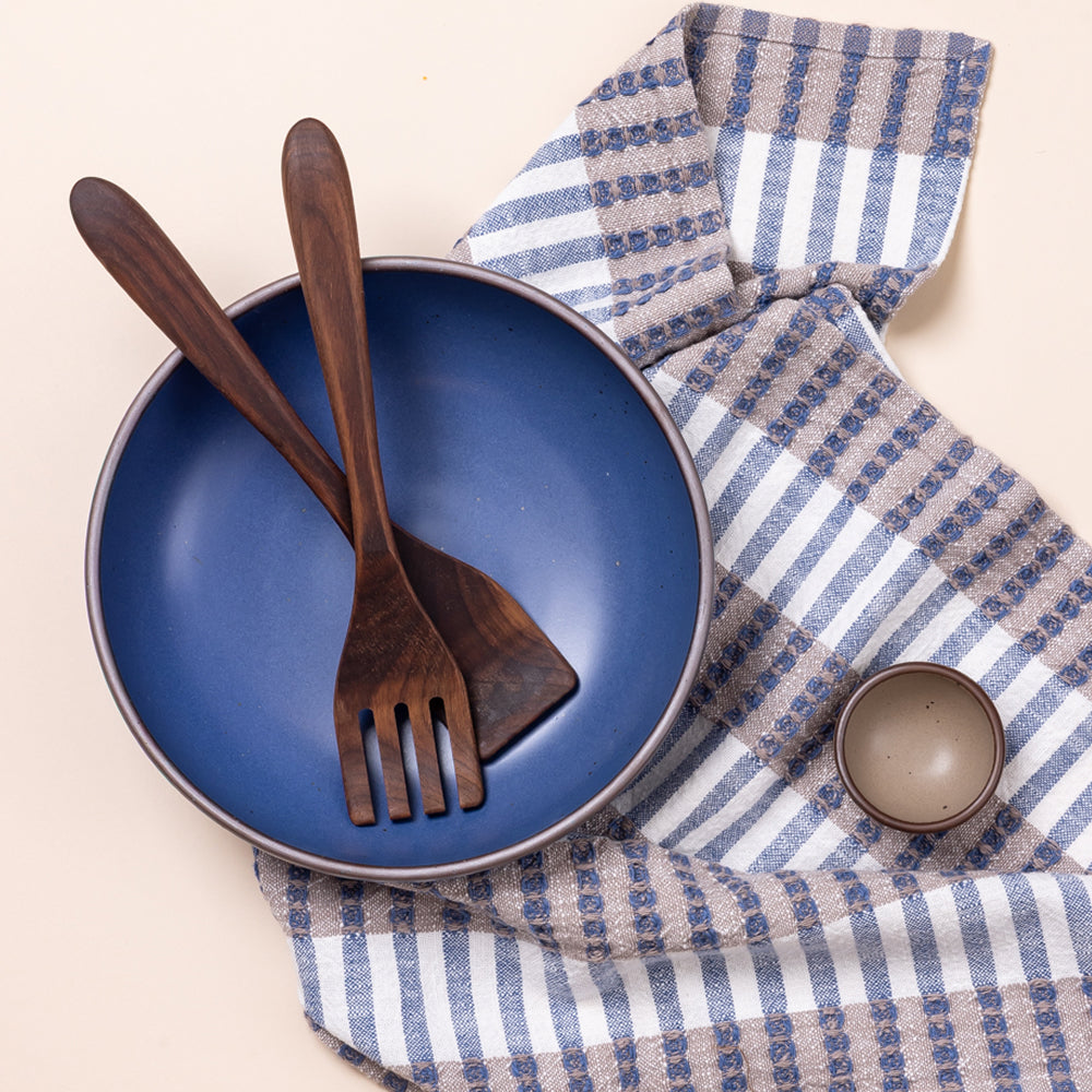 Overhead view of a large ceramic shallow serving bowl in a grounded blue color with wood serving set and striped neutral and blue tea towel.