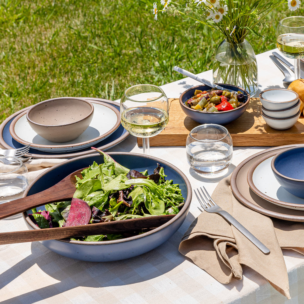 Outdoor table set with ceramic bowls and plates, salads, wine glasses, flatware, and a vase of flowers.