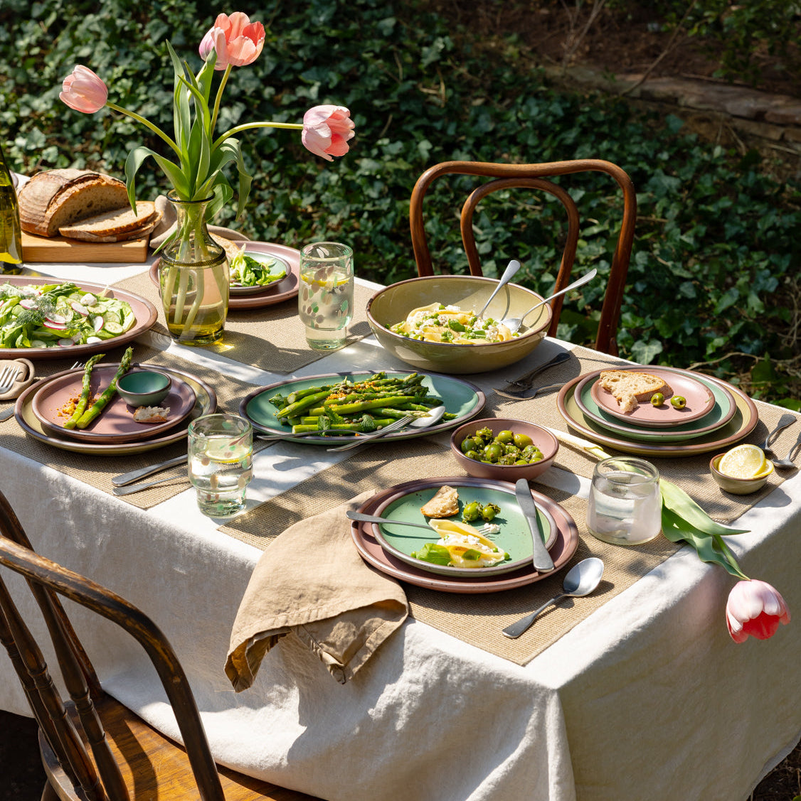 Dining table set with green and pink ceramic dinnerware, glasses, and utensils outdoors with greenery in the background.