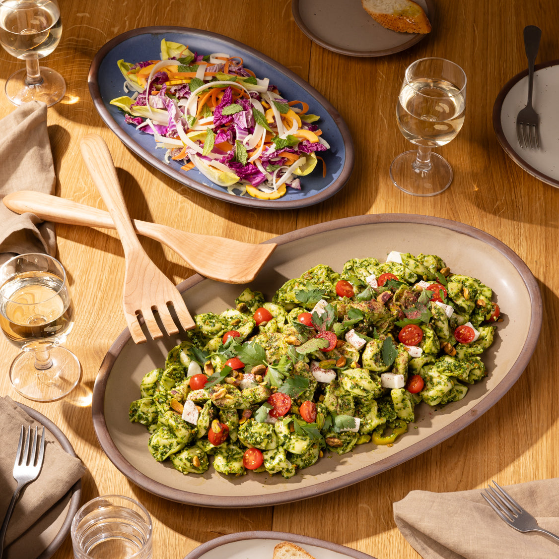 Family-style table setting with pesto pasta, colorful salad, and drinks served on oval ceramic platters and plates.