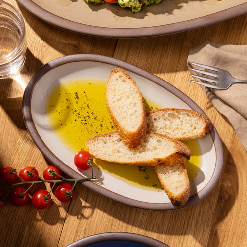 Two slices of bread with olive oil on an oval plate, surrounded by cherry tomatoes and a fork, on a wooden table.