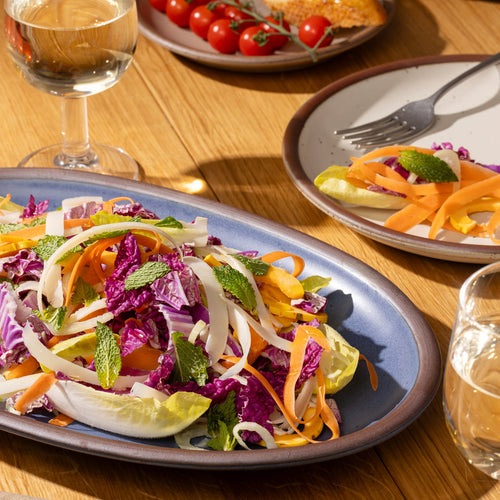 Colorful salad on a blue medium oval platter with a glass of water and a side of bread and tomatoes on a wooden table.