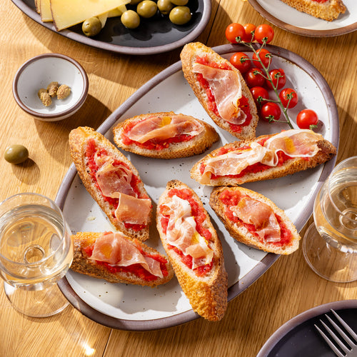 Medium white oval platter with bread, prosciutto, tomatoes, and olives on a wooden table.