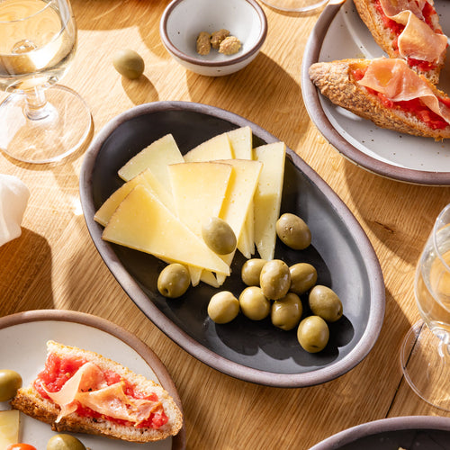 Small black ceramic oval platter of appetizers including cheese, olives, and bread on a wooden table.