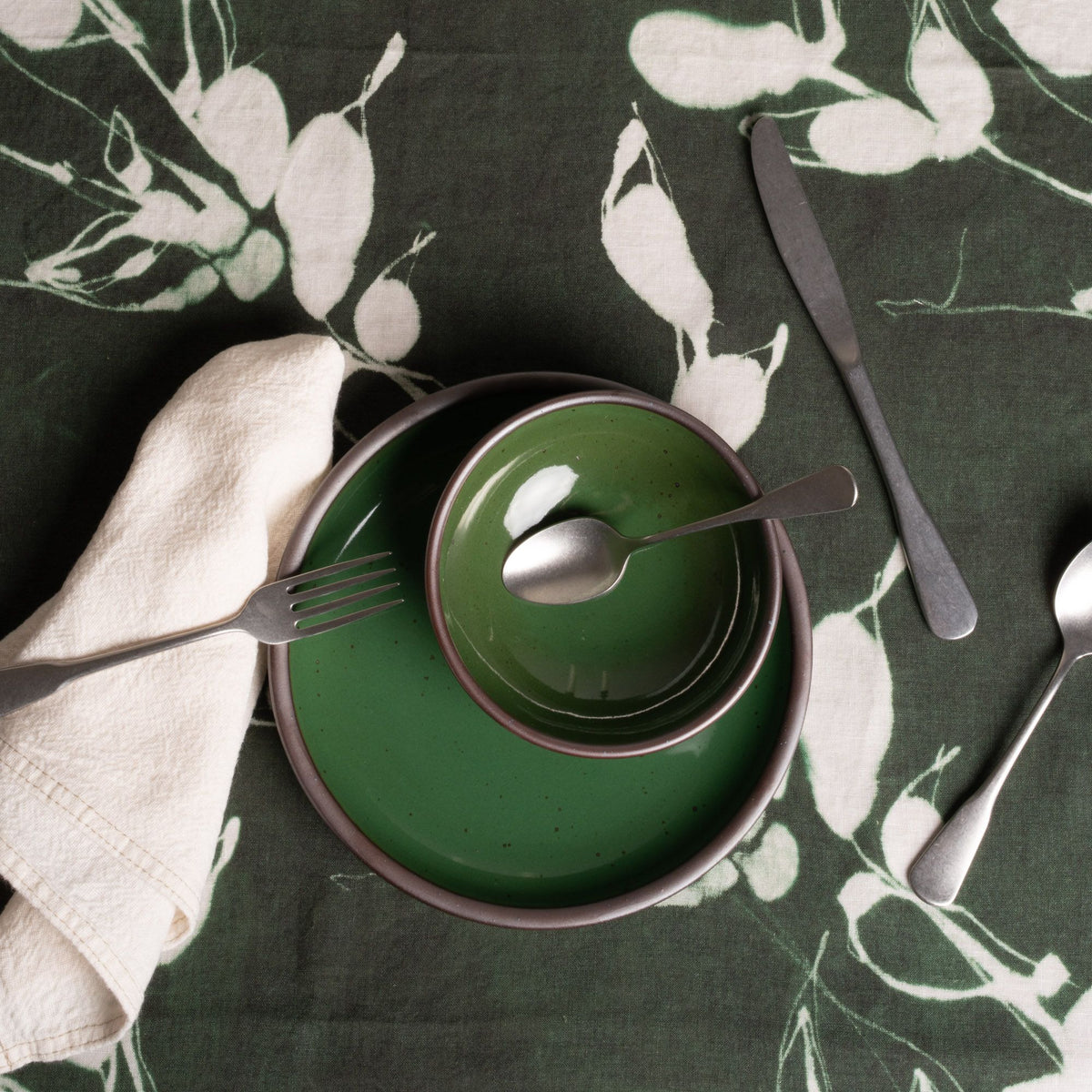 Place setting on dark green floral tablecloth with ceramic dishes, silverware, glass, and a folded cloth napkin.