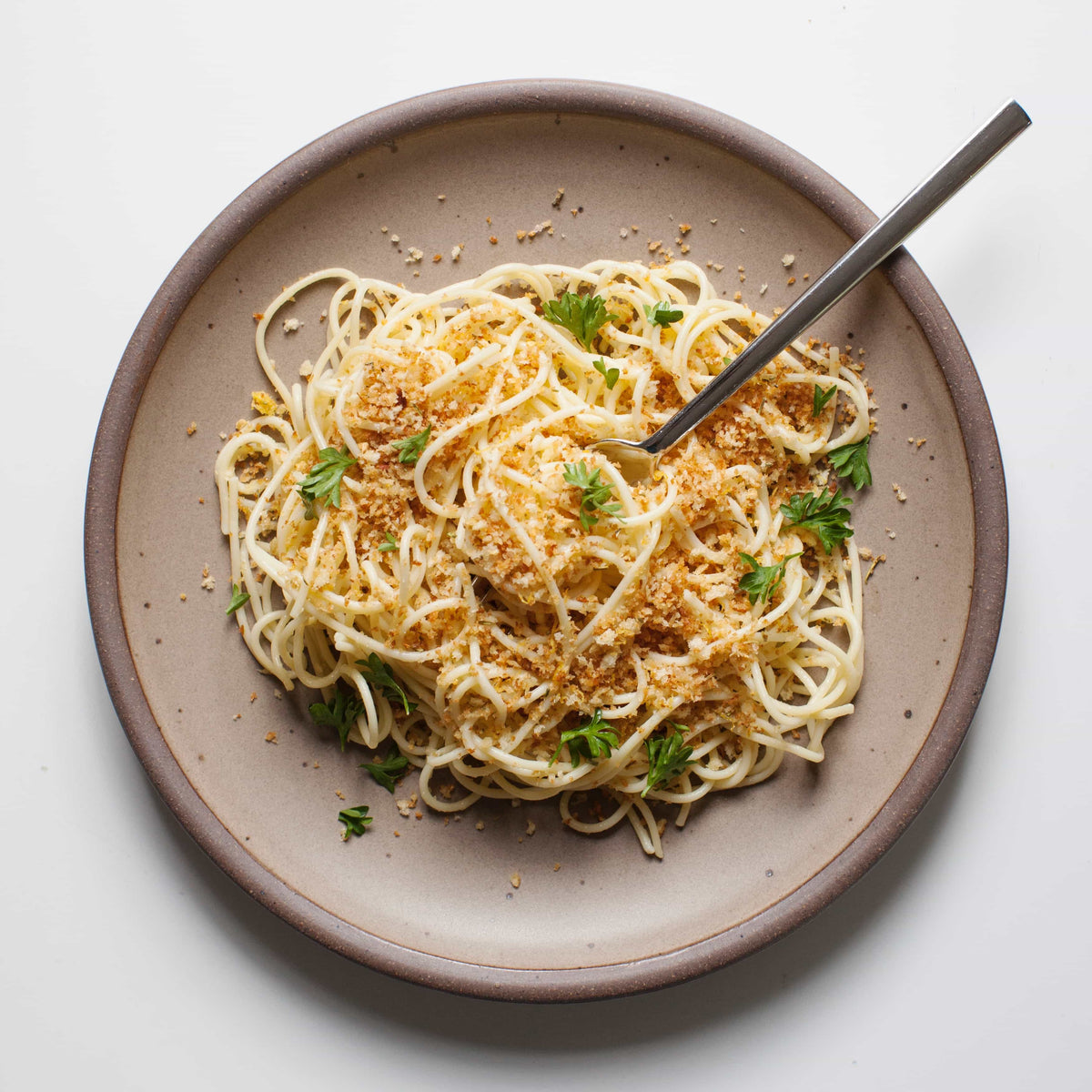 A ceramic dinner plate in a warm pale brown color featuring iron speckles and an unglazed rim with pasta on top and a fork