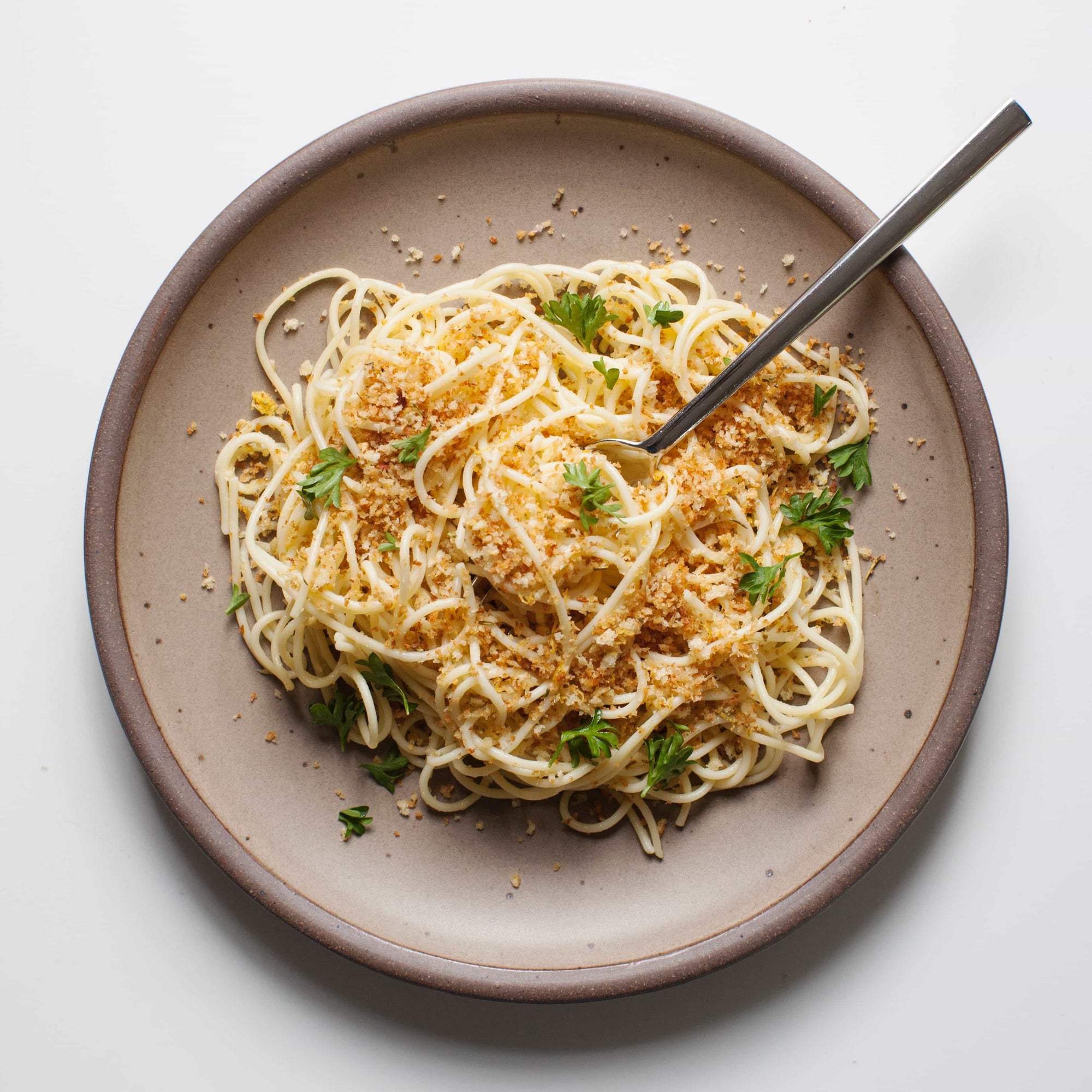 A ceramic dinner plate in a warm pale brown color featuring iron speckles and an unglazed rim with pasta on top and a fork