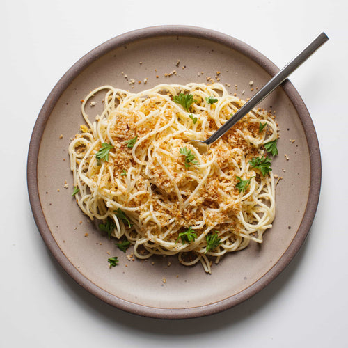 A ceramic dinner plate in a warm pale brown color featuring iron speckles and an unglazed rim with pasta on top and a fork