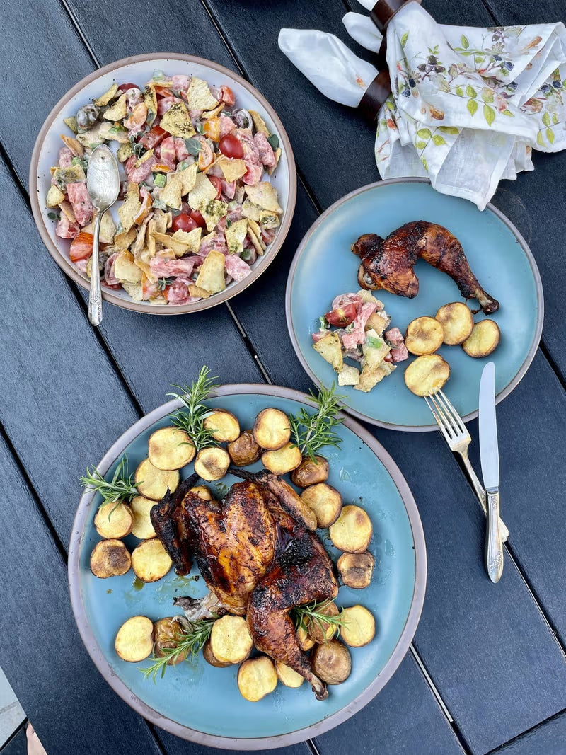 A grilled chicken surrounded by roasted potatoes and rosemary is served on a large blue plate, with a side salad on a matching plate, all set on a dark wood table with floral napkins nearby.
