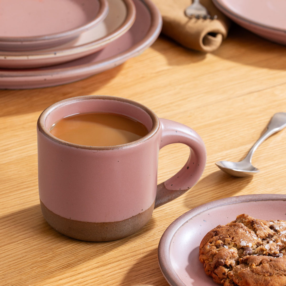 Dusty mauve pink ceramic mug filled with coffee beside a cookie on a matching plate, set on a wooden table with dishware in the background.