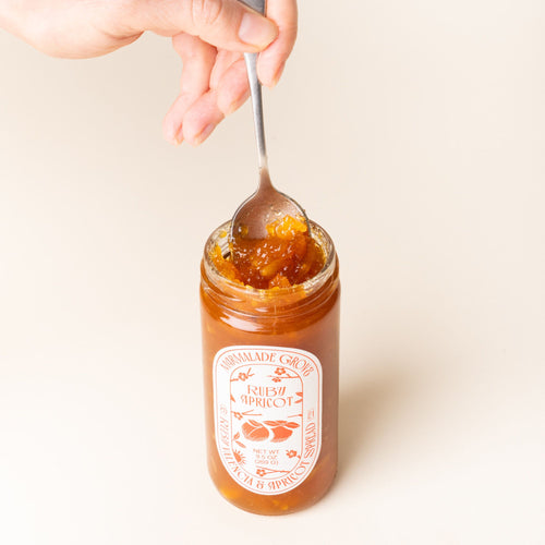 A hand holds a spoon to scoop fruit spread out of an open jar of Marmalade Grove Ruby Apricot Spread, featuring a red and white label with fruit and floral illustrations.
