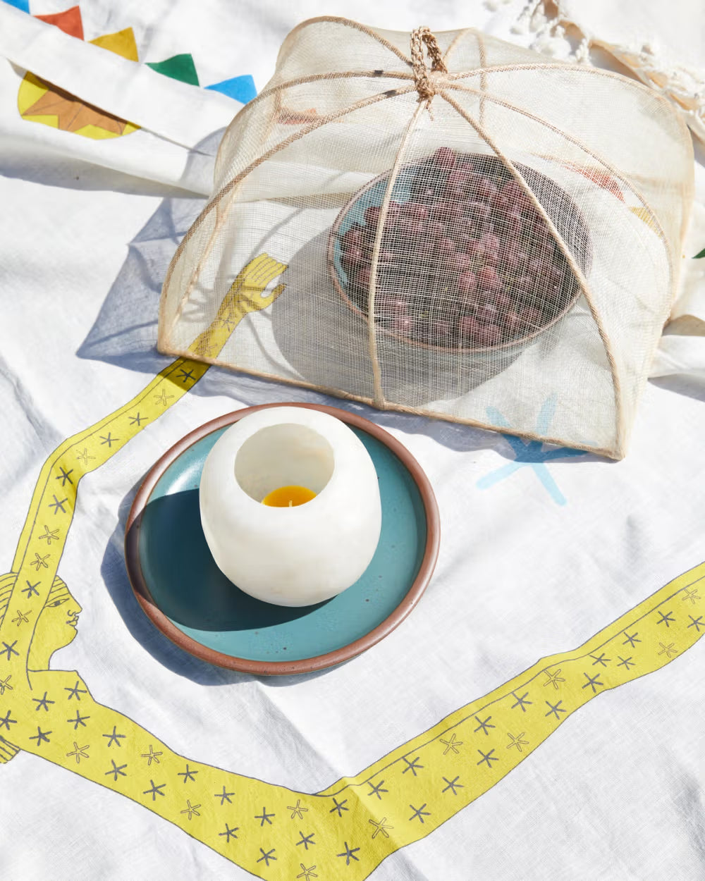 A bowl of berries covered with a mesh food cover sits on a white cloth with colorful geometric patterns. Nearby, a candle in a stone holder is placed on a turquoise plate with a brown rim.