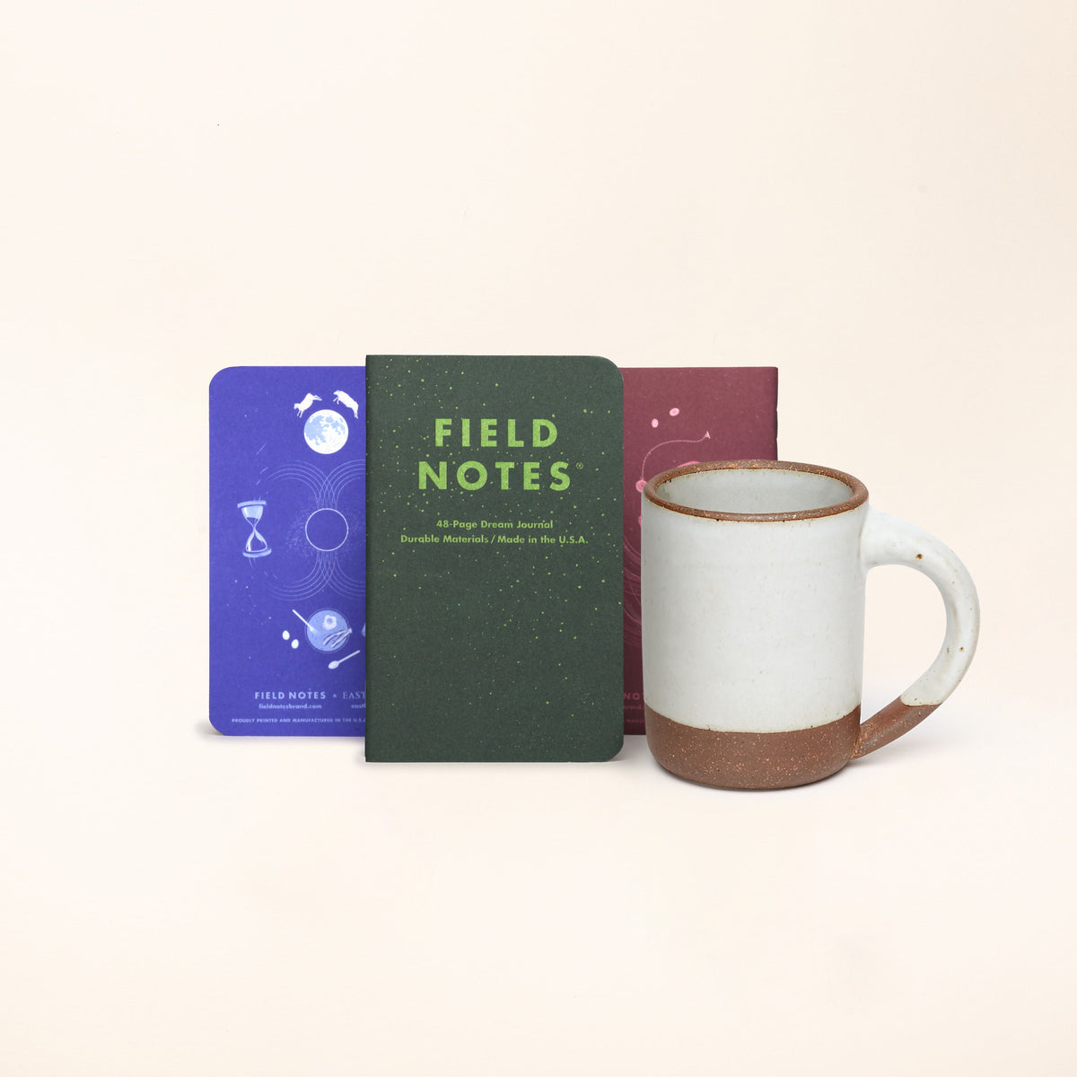A 3 pack of Field Notes Journal standing up in a row in forest green, lapis blue, and plum colors with The Mug in a cool white color.