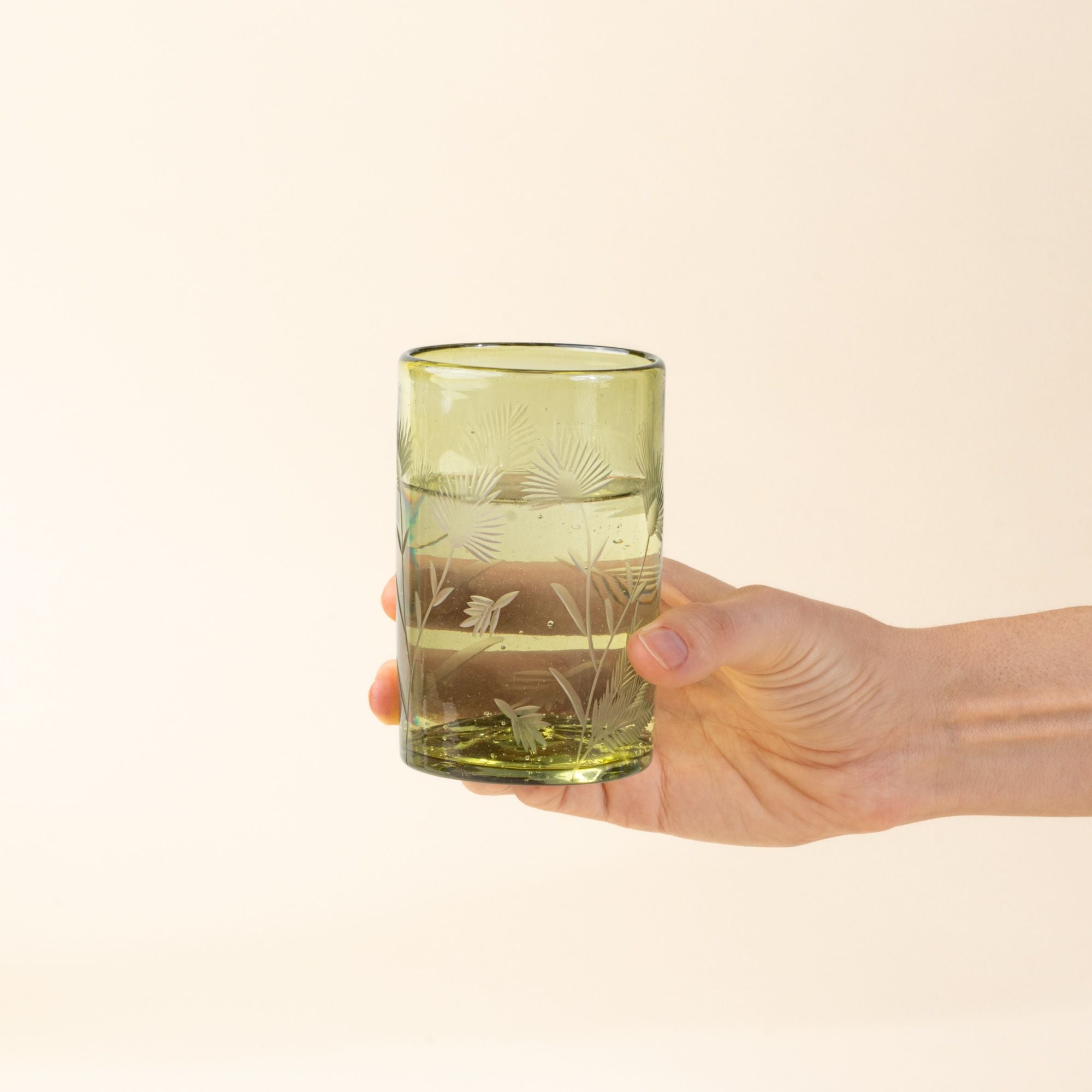 A hand holds out an olive tumbler glass made of recycled glass with etched thistle designs on it filled with water.