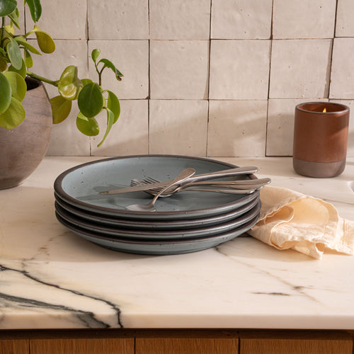 A stack of 4 dinner sized ceramic plates in a blue-grey color featuring iron speckles and an unglazed rim, with silverware and napkin on a countertop