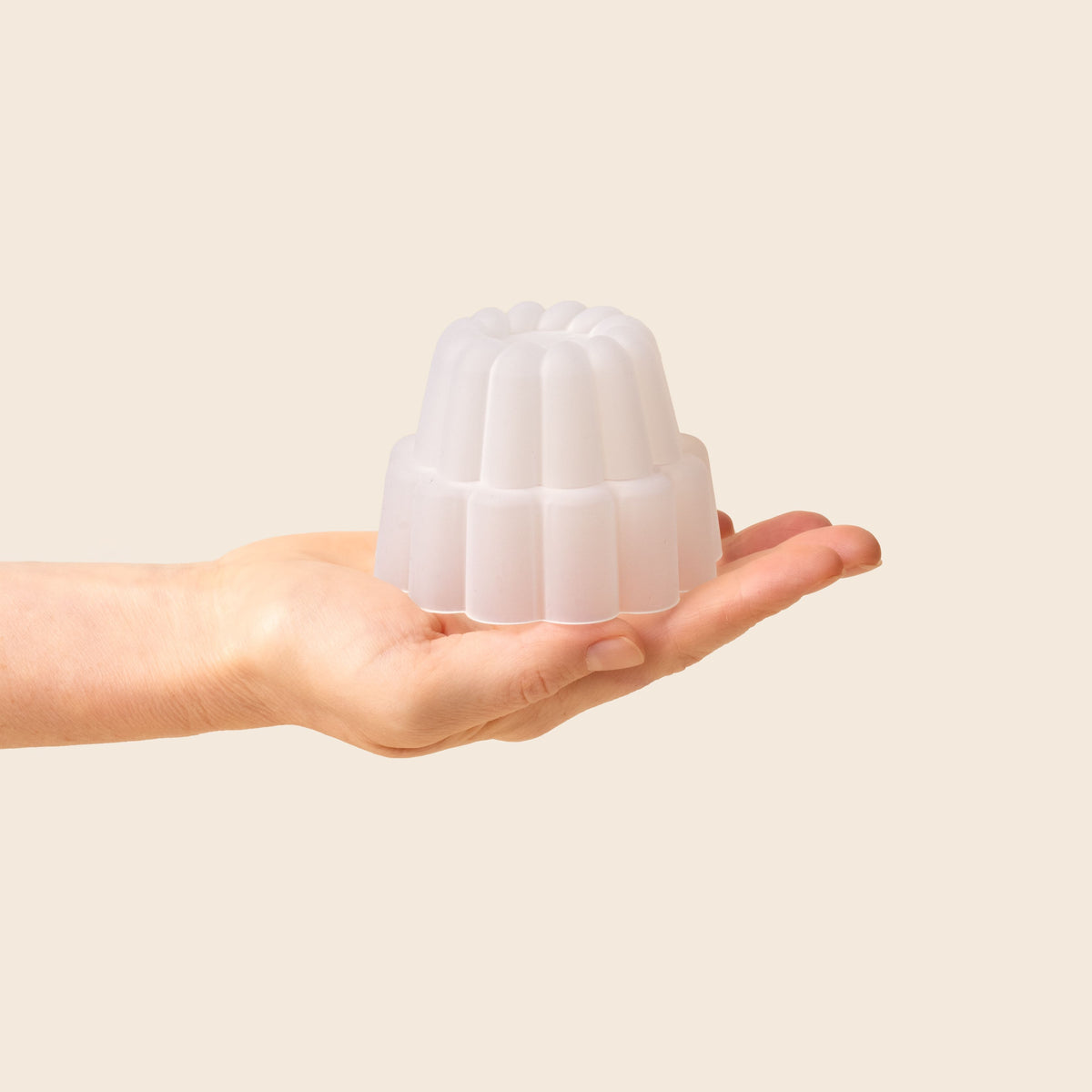 A hand holding a small, white, fluted jelly-shaped mould for dessert set against a soft beige background.