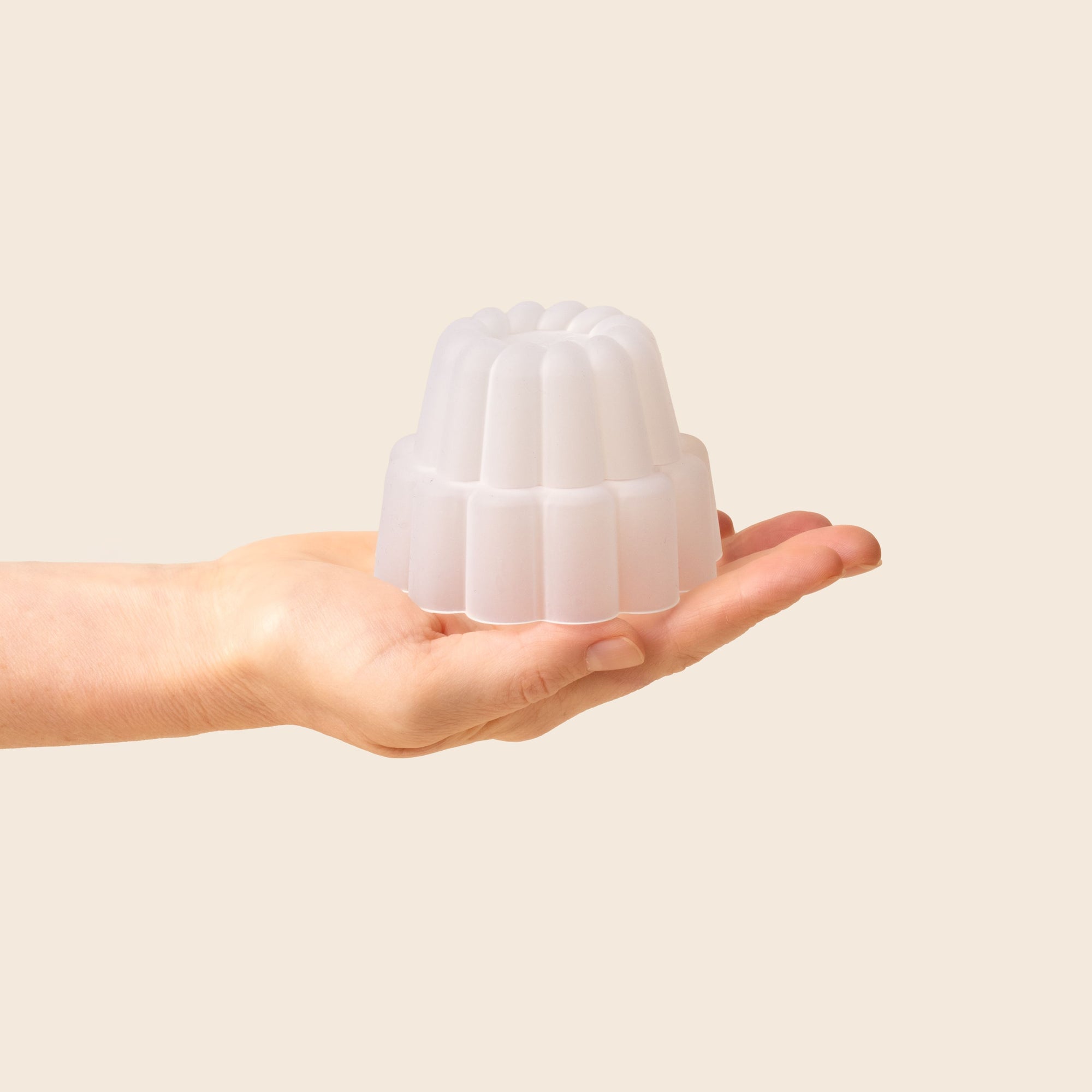 A hand holding a small, white, fluted jelly-shaped mould for dessert set against a soft beige background.