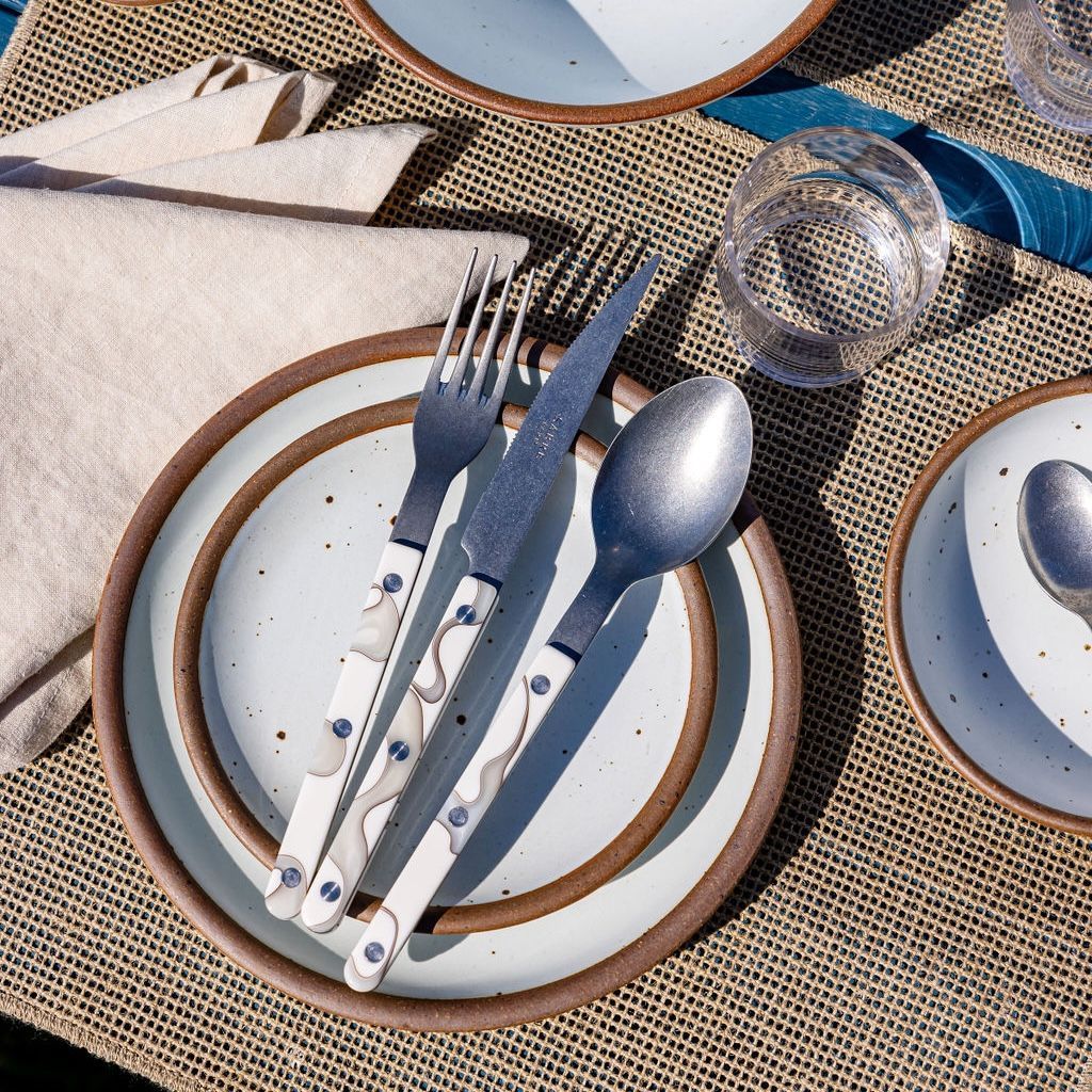 A table setting with a neutral woven placemat, stacked cool white ceramic plates, and ivory marble-handled fork, knife, and spoon.