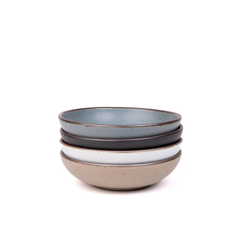 A stack of 4 dinner-sized shallow ceramic bowls in a blue-grey, graphite black, cool white, and pale warm brown colors featuring iron speckles and an unglazed rim