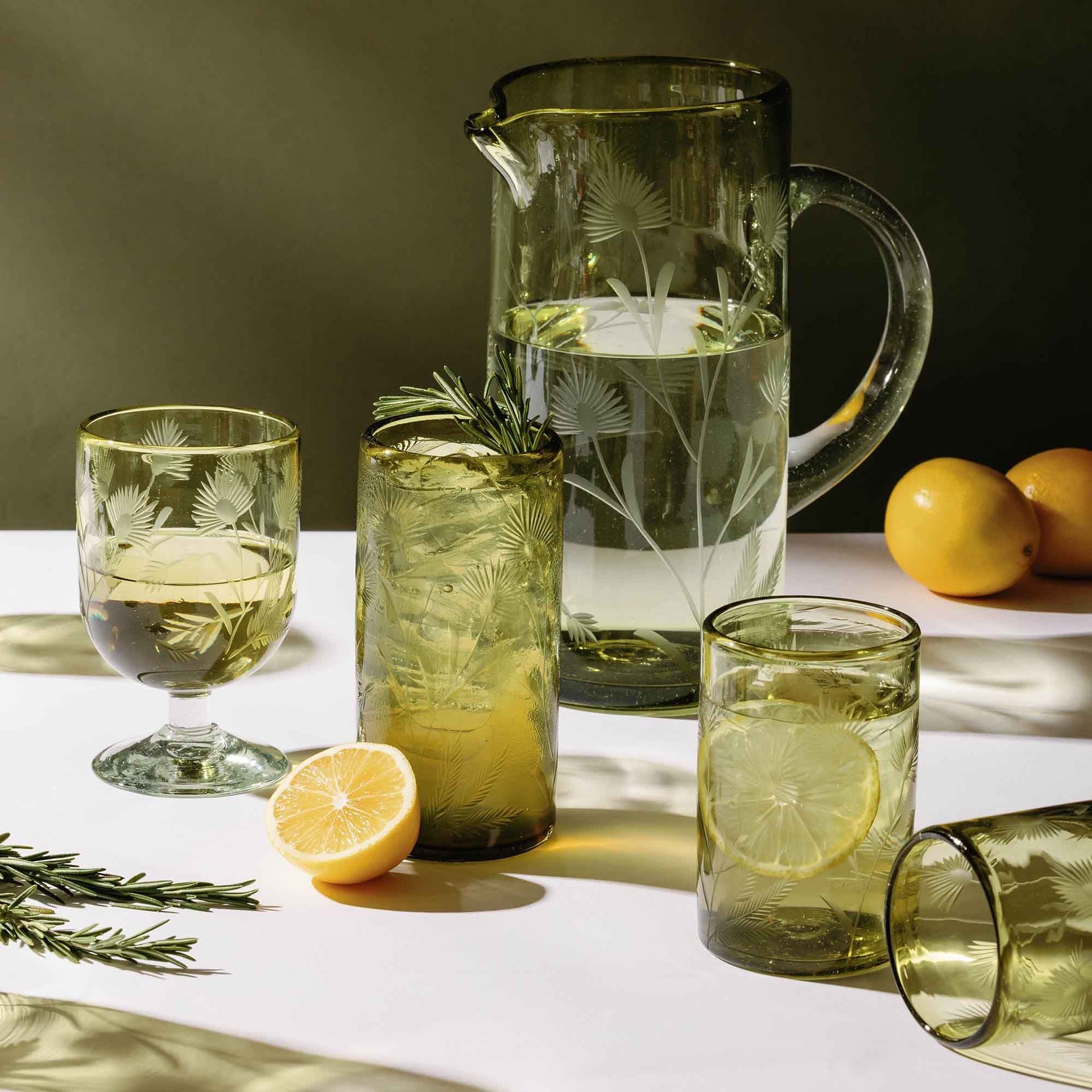 Green etched glass drinkware set with lemon slices and rosemary on a sunlit table.