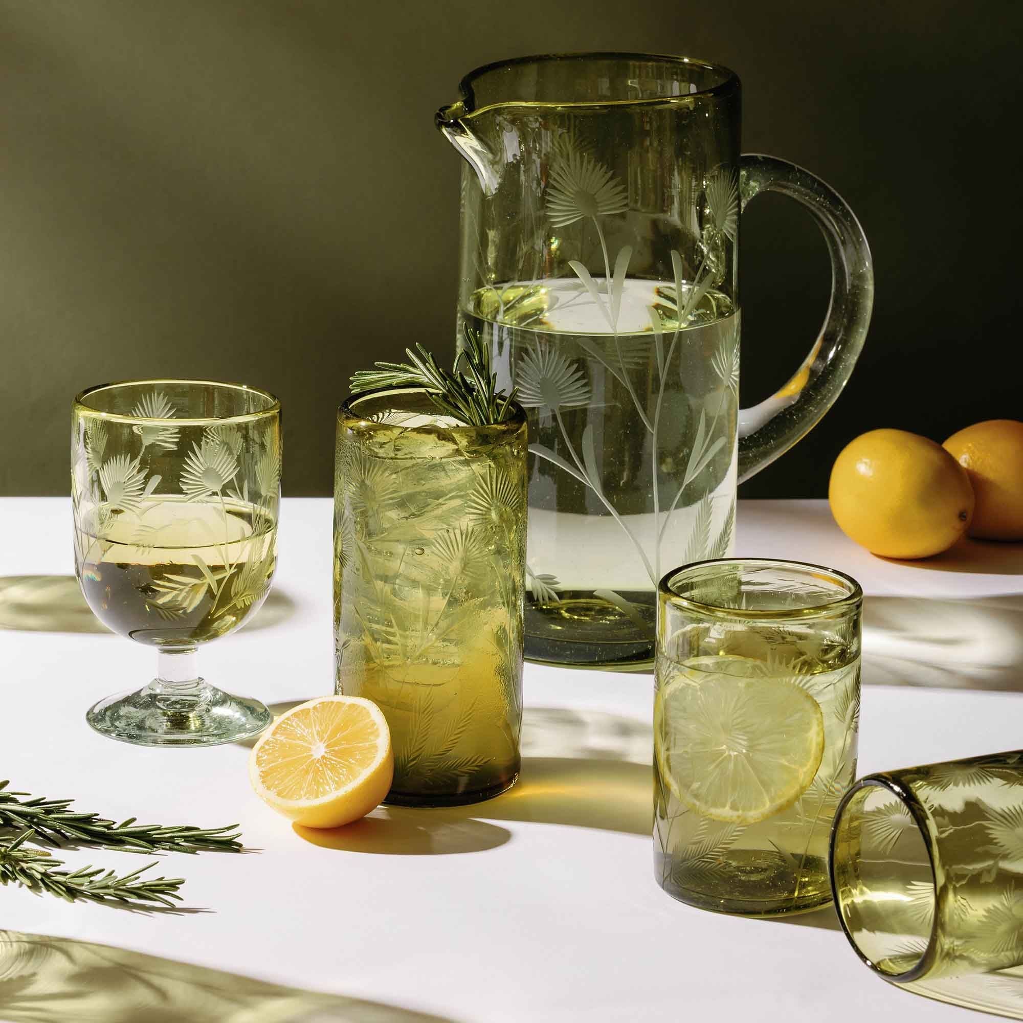 Green etched glass drinkware set with lemon slices and rosemary on a sunlit table.
