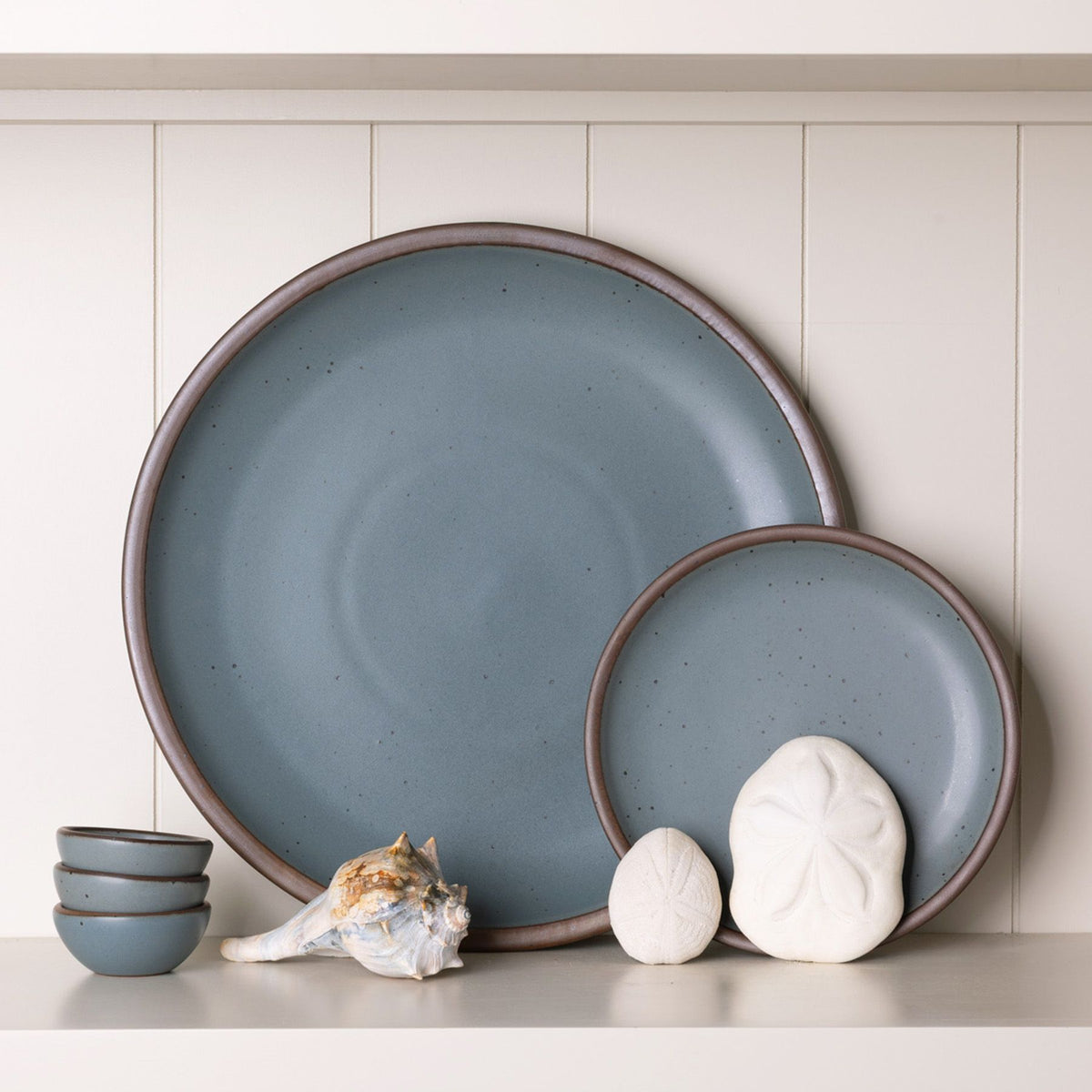 A large ceramic serving platter and side plate in a blue-grey color propped on a shelf with shells and a stack of small bowls in the same color.