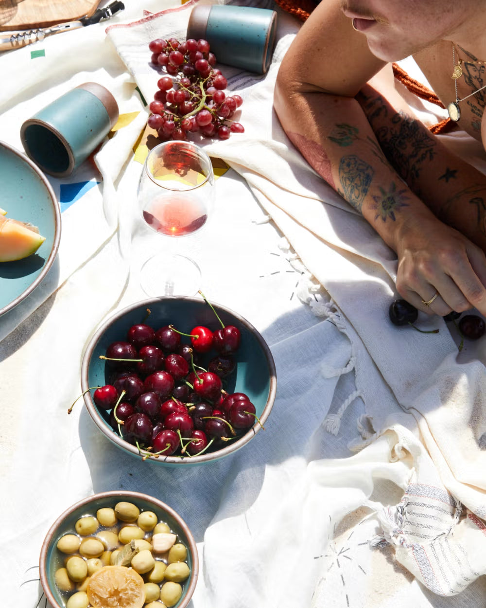 A sunlit picnic scene with a person lying on a patterned blanket, surrounded by ceramic bowls filled with cherries and olives. A glass of rosé, a small cluster of red grapes, and teal ceramic tumblers are scattered nearby.
