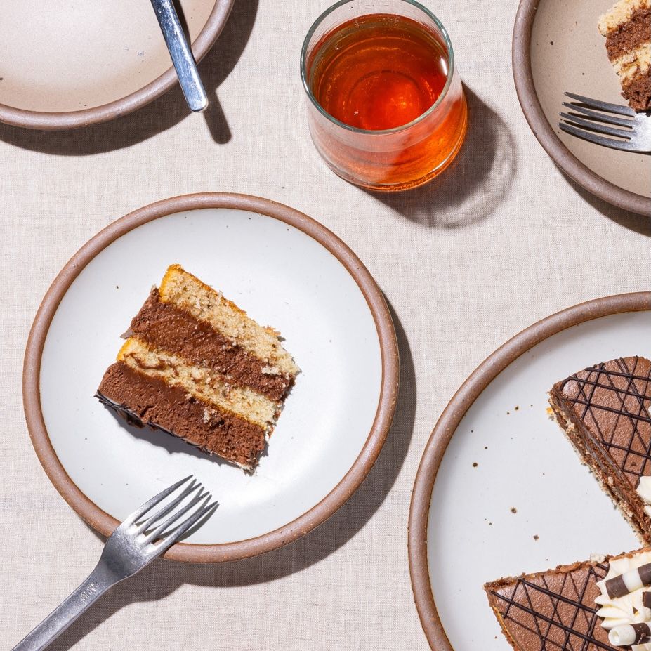 Plates of various sizes in white and a muted light brown are filled with small slices of cake, silver forks, and a glass of red juice.