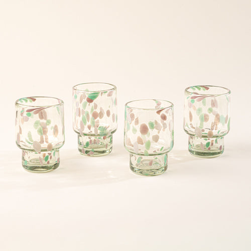 Four short clear glasses with pastel green and pink speckled patterns arranged in a row on a light background.