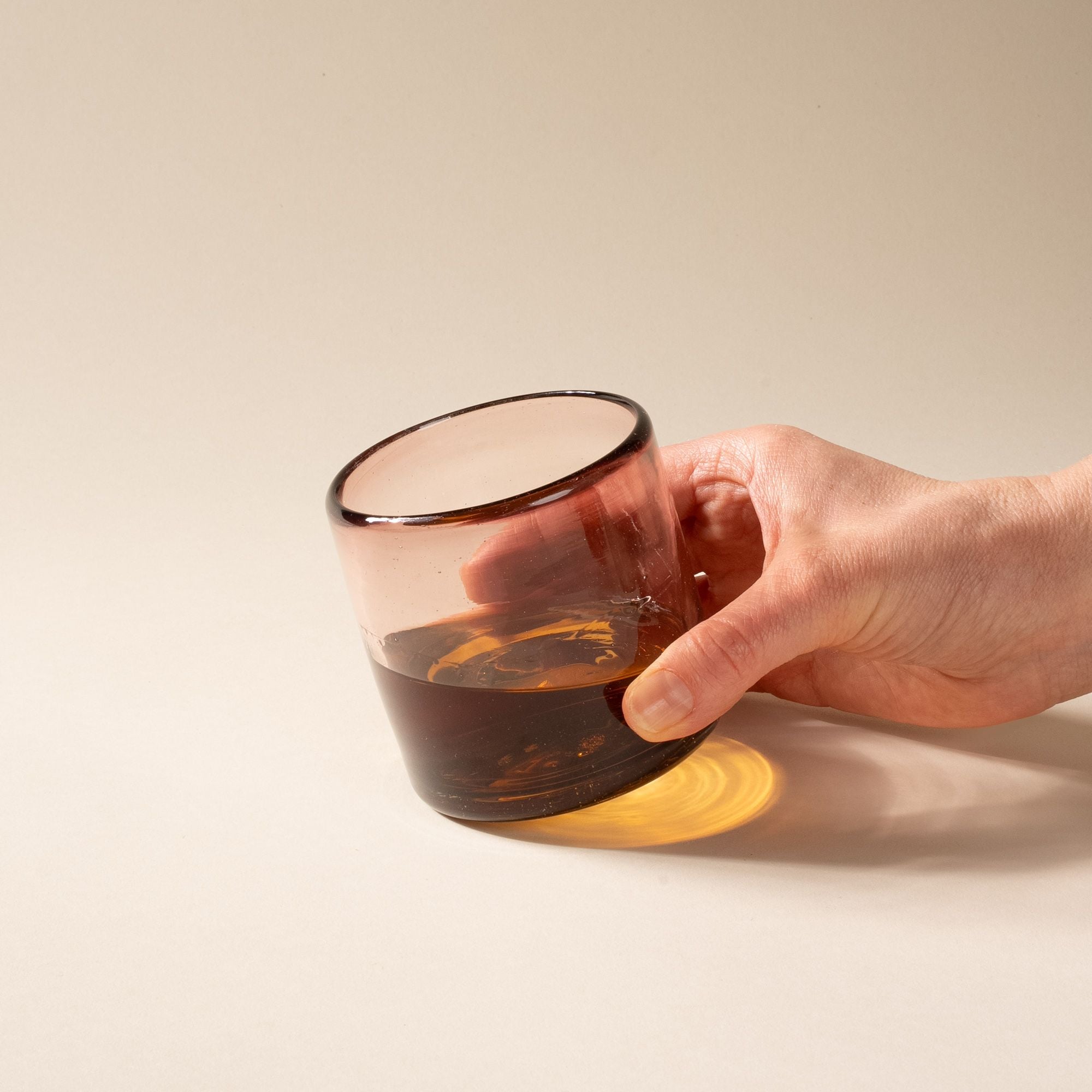 A hand holding a light plum short transparent drinking glass with whiskey inside