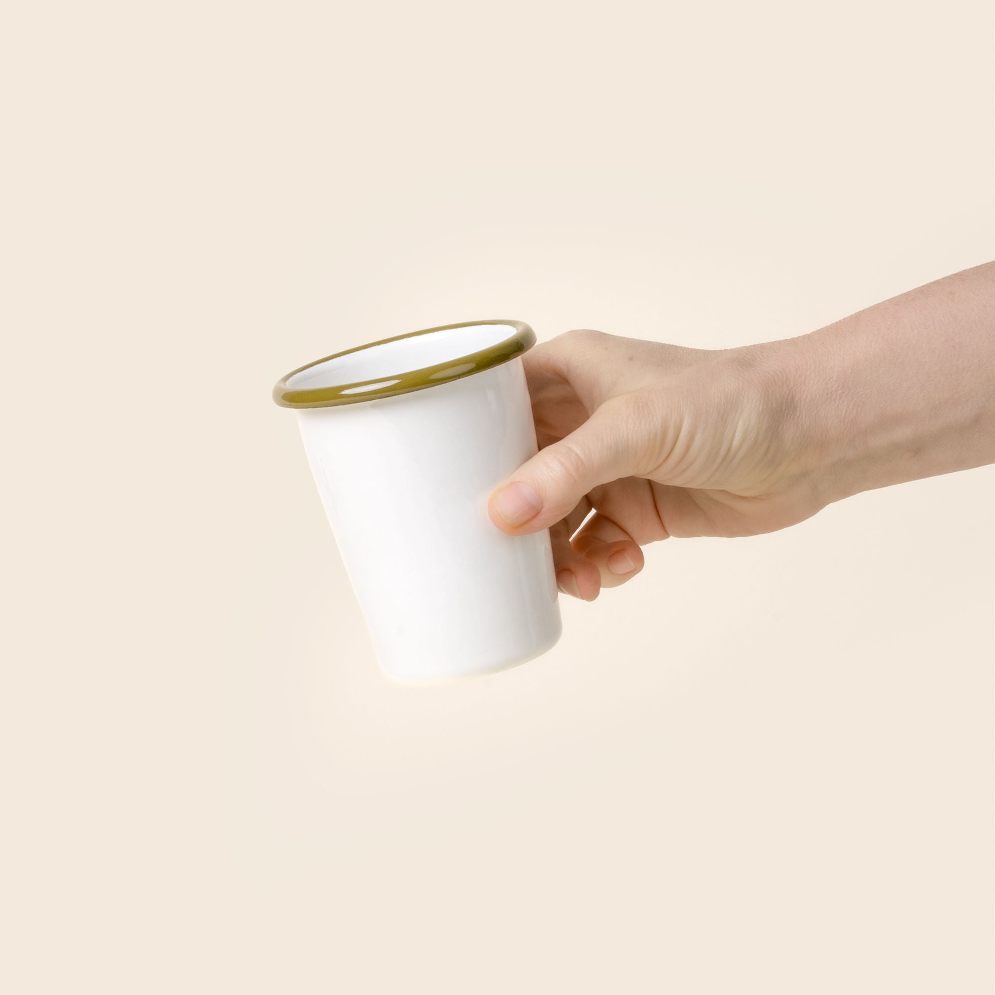 A hand holds a short white enamel tumbler with an olive green rim, set against a soft beige background.