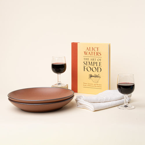A copy of The Art of Simple Food by Alice Waters sits beside two glasses of red wine, stacked plates, and folded white napkins.