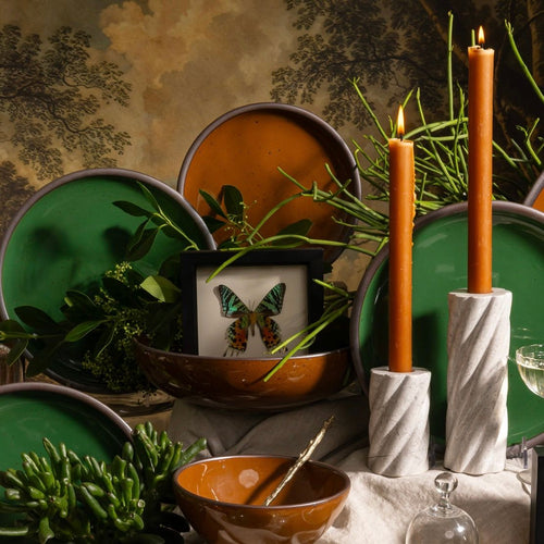 Glossy green and brown ceramic plates with greenery, lit terracotta taper candles in marble holders, and framed butterfly decor.