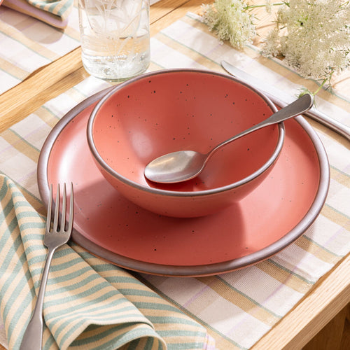 Watermelon pink ceramic soup bowl and plate set on a checkered placemat with a striped napkin, silverware, and etched glass.