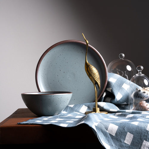 A round ceramic bowl and propped dinner plate in a blue-grey color, with an artfully arranged brass bird and crosshatch tablecloth
