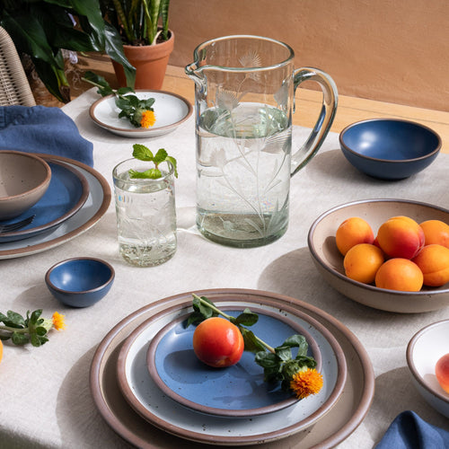 An etched thistle tumbler glass and pitcher filled with water, on a table with ceramic dinnerware and fruit, with plants in the background.